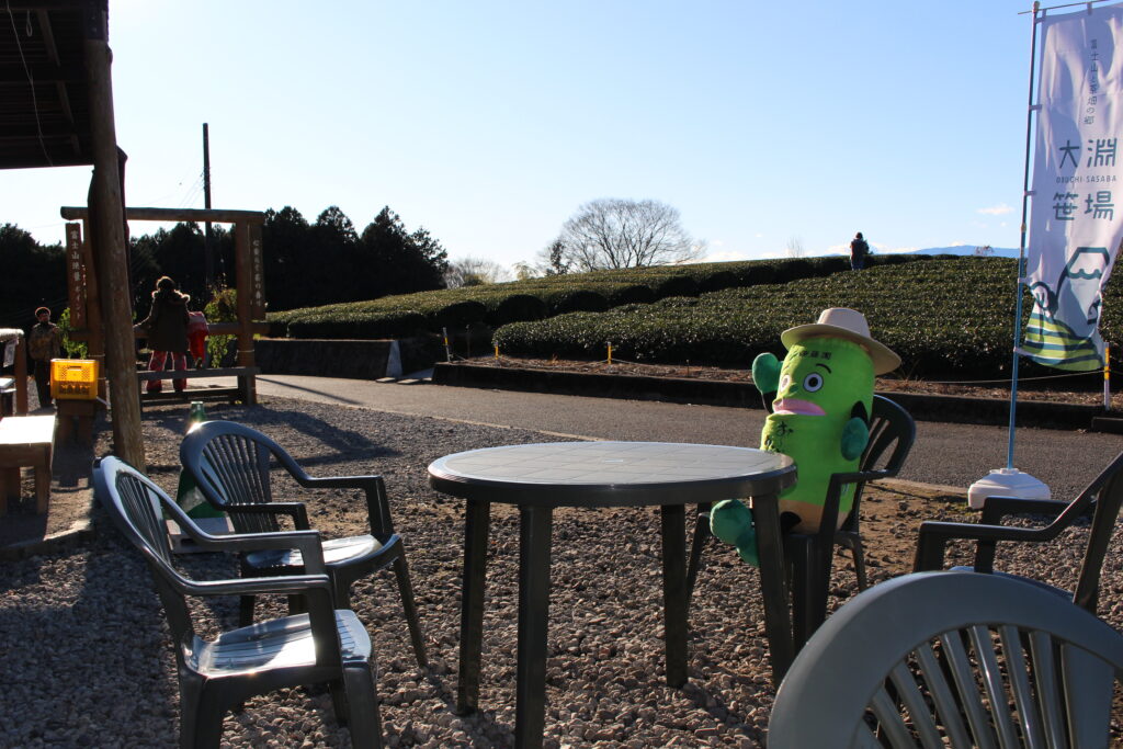 大淵笹場の休憩スペースにあるテラス席でくつろぐ、「お〜いお茶」の公式キャラクター「お〜いお茶くん」。茶畑を眺めながら一息つける現地の雰囲気を紹介。