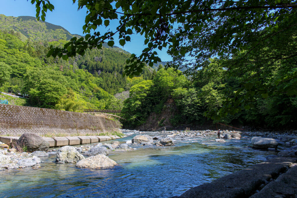 青空のもと、新緑に彩られた山々と透明度の高い道志川の清流。WOODSMAN CAMPGROUNDのテントサイトの目の前に広がる開放的な大自然。