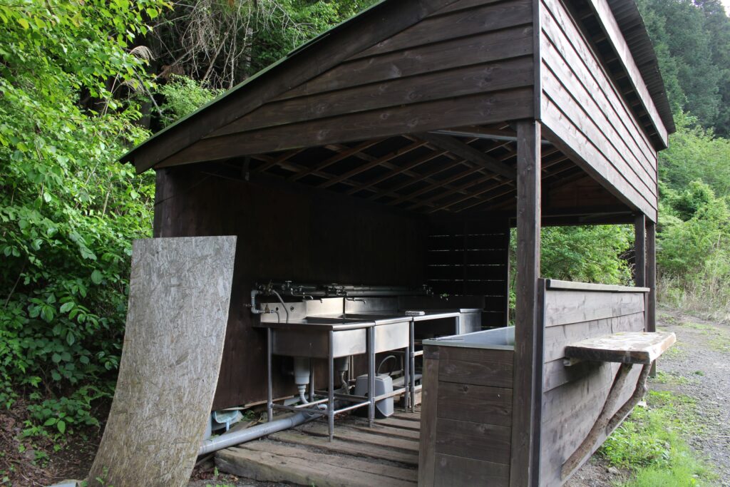 WOODSMAN CAMPGROUNDの、周囲の自然に溶け込む木造の屋根付き炊事場。清潔に保たれたステンレス製のシンクと、森の緑が背景に広がる。