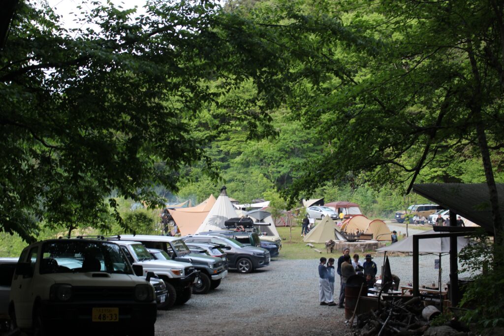 WOODSMAN CAMPGROUNDの木々の隙間から見下ろす、様々なテントが並ぶ賑やかなキャンプサイトと、手前に並ぶキャンパーたちの車。