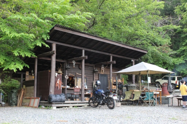 山梨のWOODSMAN CAMPGROUNDにある木造の渋い管理棟と、その前に美しく停められたネイキッドバイク。豊かな緑に囲まれた隠れ家のような受付エリア。