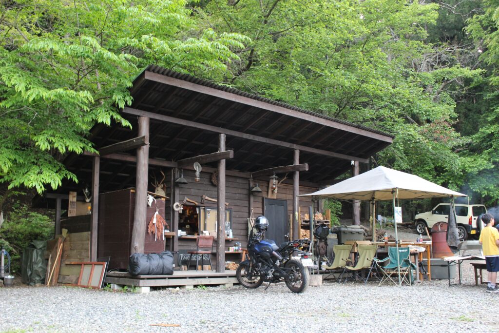 山梨のWOODSMAN CAMPGROUNDにある木造の渋い管理棟と、その前に美しく停められたネイキッドバイク。豊かな緑に囲まれた隠れ家のような受付エリア。