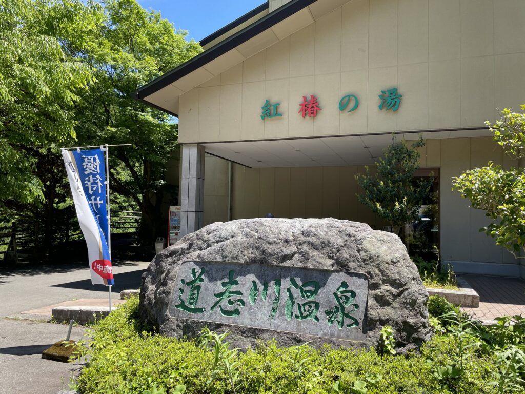 道志川温泉紅椿の湯の入り口に佇む立派な石碑と新緑の景色。山梨県道志村でのソロキャンプやツーリング帰りに立ち寄る、極上の日帰り温泉の雰囲気を伝えるエントランス。