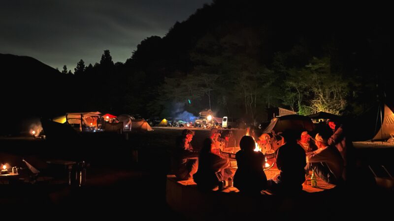 WOODSMAN CAMPGROUNDのメインサイトで、常備された大きな焚き火場を囲んで団らんする人々。夜の静寂の中に広がるキャンプファイヤーの温かい雰囲気。