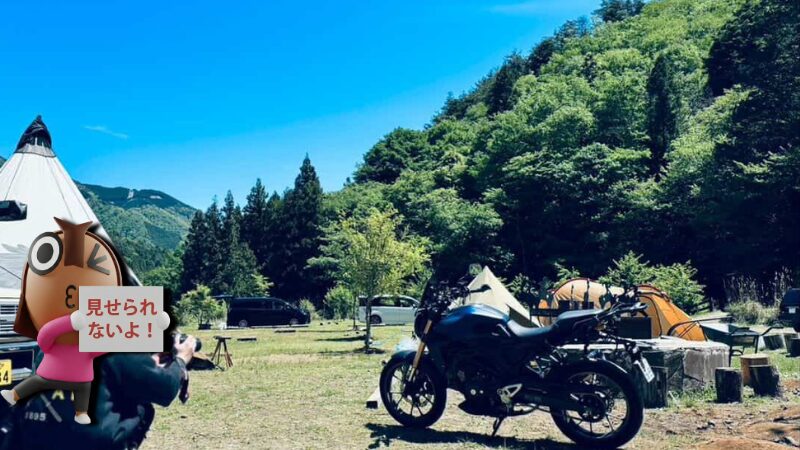 快晴のWOODSMAN CAMPGROUNDメイン広場で愛車CB250Rをカメラで撮影するねりすけ。豊かな緑に囲まれた芝生サイトで、バイクキャンプツーリングの思い出を切り取る瞬間。