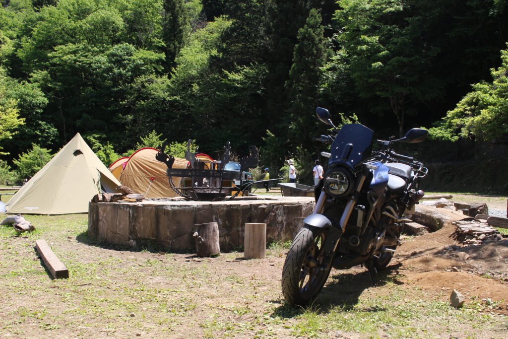 WOODSMAN CAMPGROUNDのメインサイトに佇む愛車CB250R。特注のファイヤーピットやテントを背景に、道志の豊かな自然の中で楽しむバイクキャンプのロケーション実例。
