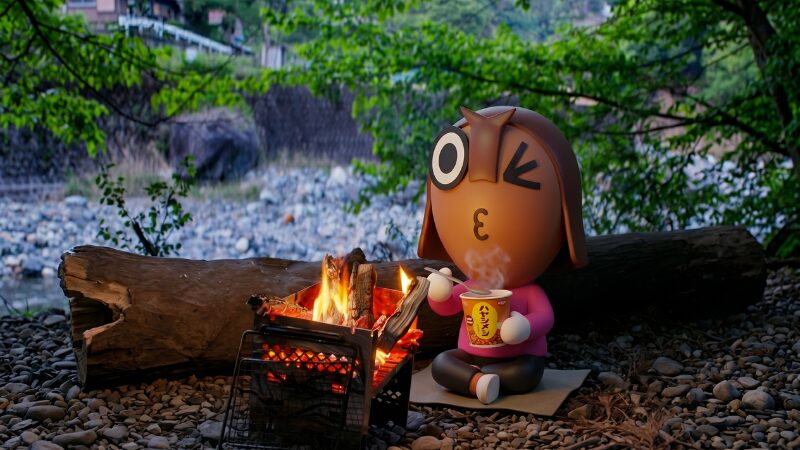 山梨県道志のWOODSMAN CAMPGROUNDの河原で焚き火を囲み、日清のハヤシメシを食べるねりすけ。道志川の豊かな自然の中で楽しむ、簡単で温かいキャンプ朝食の体験実例。