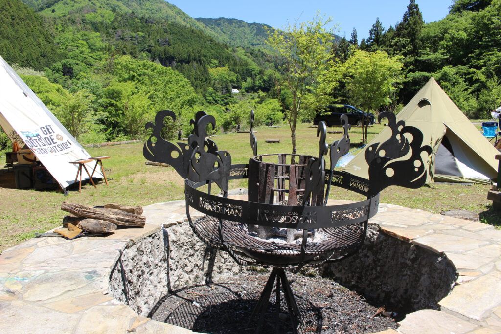 WOODSMAN CAMPGROUNDのメインサイト中央に常設された石組みの大型ファイヤーピット。ショップロゴが刻まれたアイアン製の特注焚き火台がある共有スペース。